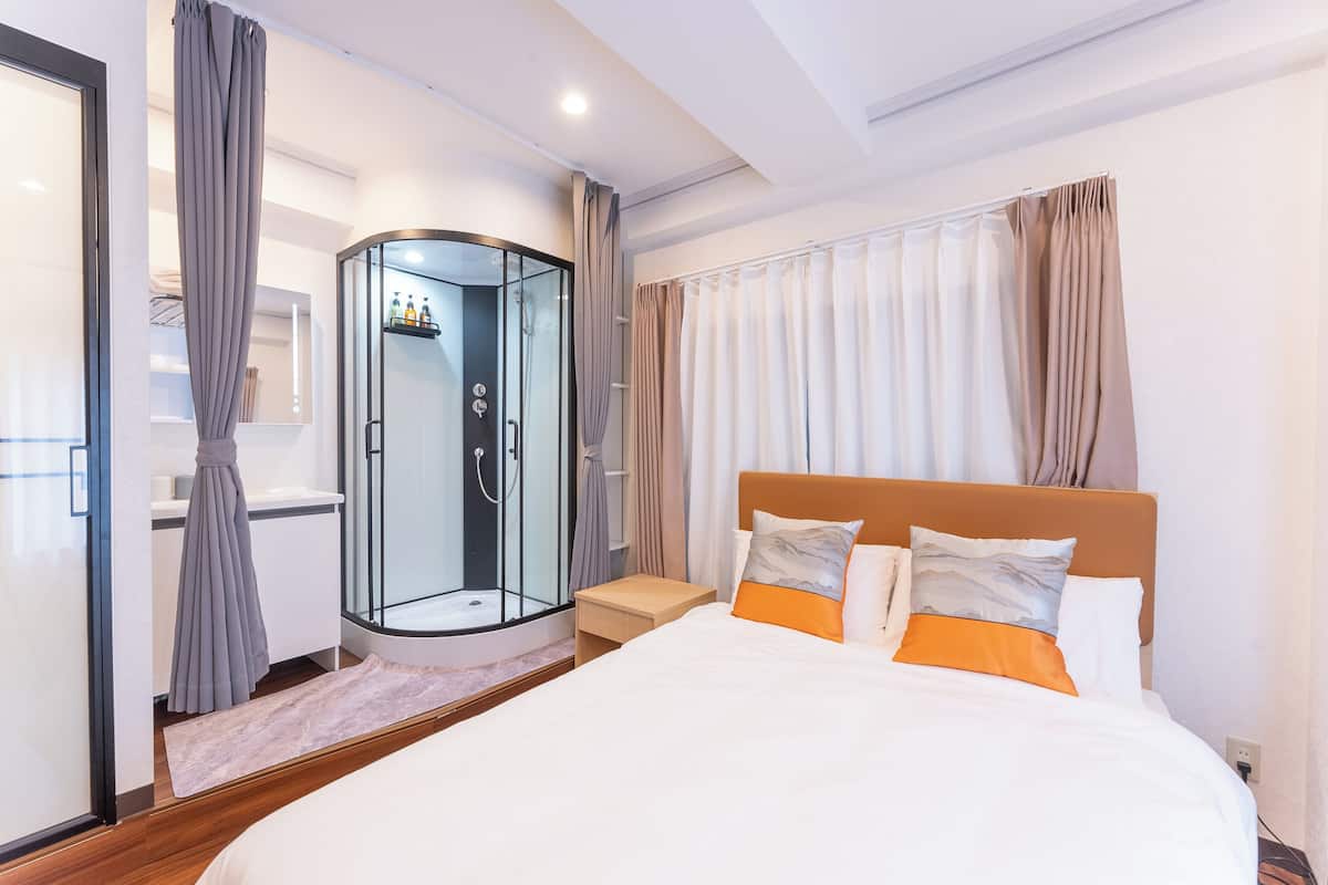 Quarto de hotel moderno com uma cama grande, chuveiro em vidro e mesa de cabeceira de madeira.