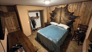 A bedroom with a large wooden headboard, a bed with blue bedding, a wooden dresser, a chair, and a coat rack with clothes. - Enchanting cabin on the river in charming Anchor Point, AK (Anchor Point)