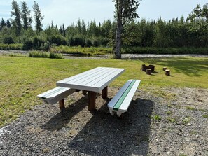 A picnic table with benches in a grassy outdoor area with trees in the background. - Enchanting cabin on the river in charming Anchor Point, AK (Anchor Point)