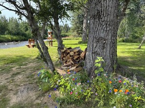 A rustic outdoor setting with a wooden bench, a small table, and a stack of firewood under a tree. - Enchanting cabin on the river in charming Anchor Point, AK (Anchor Point)