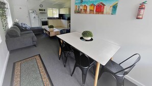 A modern kitchen with white cabinets, a dining area with a white table and black chairs, and a painting of colorful houses on the wall. - Esperance Little Units 1 (Castletown)