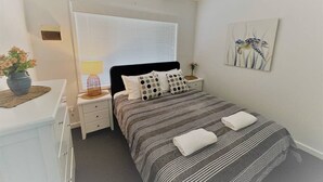 A bedroom with a bed, bedside tables, a nightstand, a window with blinds, a wall-mounted artwork, and a vase with flowers. - Esperance Little Units 1 (Castletown)