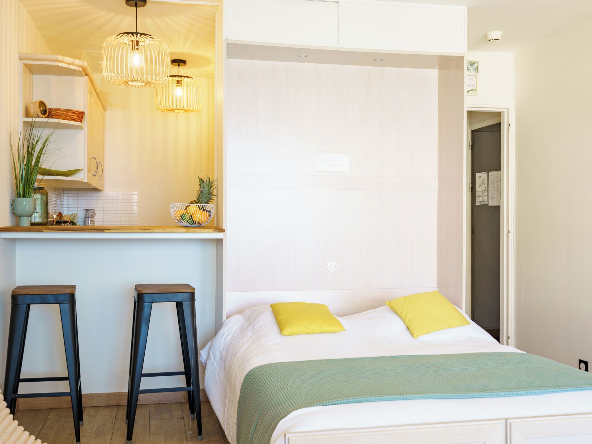 A modern hotel room with a bed, two stools, a small kitchen area, and a door leading to another room.