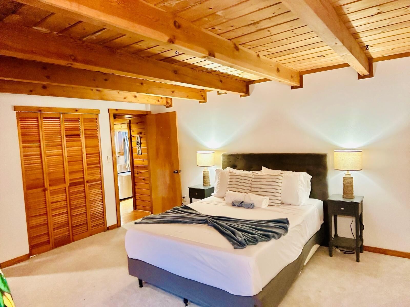 Een slaapkamer met een houten plafond, een bed met wit beddengoed, twee bedlampjes en een kledingkast met schuifdeuren.