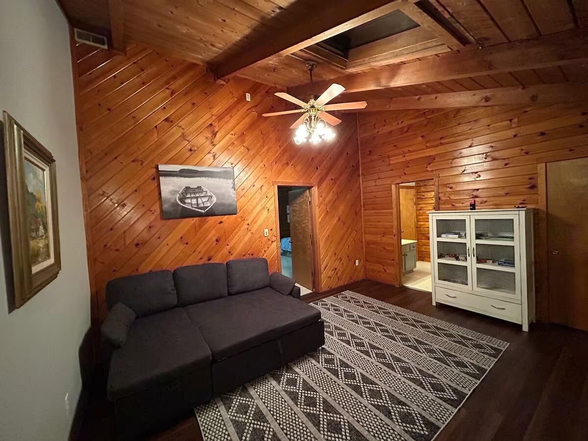 A cozy room with wooden walls, a ceiling fan, a dark sofa, a patterned rug, and a white cabinet.
