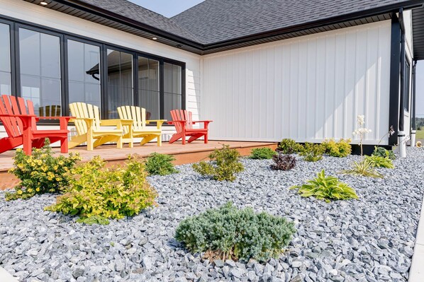 A modern house with a patio area featuring red and yellow Adirondack chairs, a small garden with various plants, and a large window overlooking the garden.