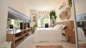 A modern bedroom with a large bed, a TV, a desk, and a window with a view of greenery.