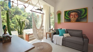 A living room with a hammock chair, a sofa, a dining table, and a view of greenery outside.