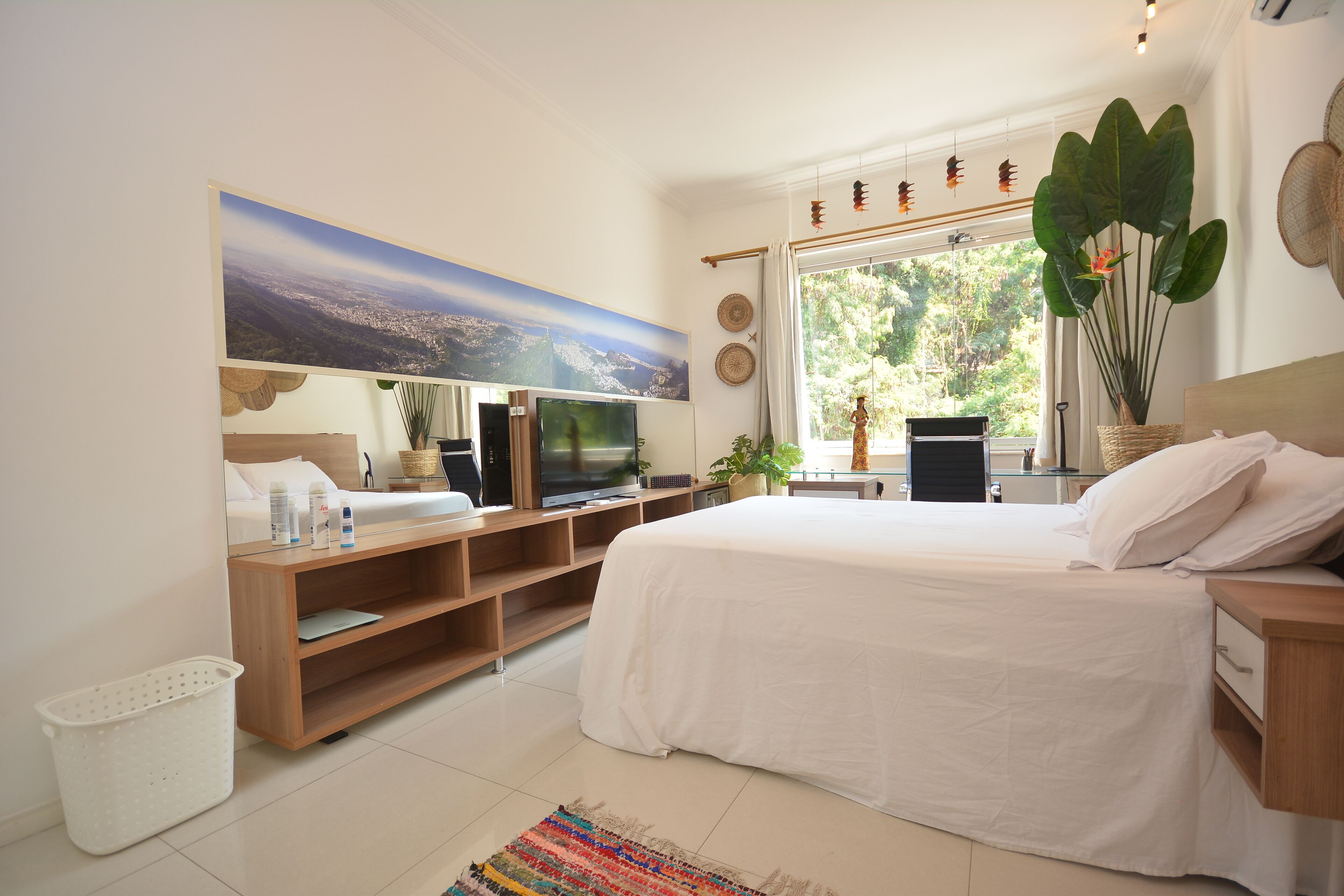 A modern bedroom with a large bed, a flat-screen TV, a wooden TV stand, a large window with a view of trees, and a wall-mounted artwork.