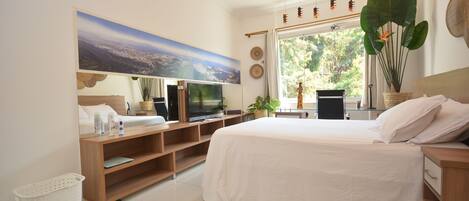 A modern bedroom with a large bed, a flat-screen TV, a wooden TV stand, a large window with a view of trees, and a wall-mounted artwork.