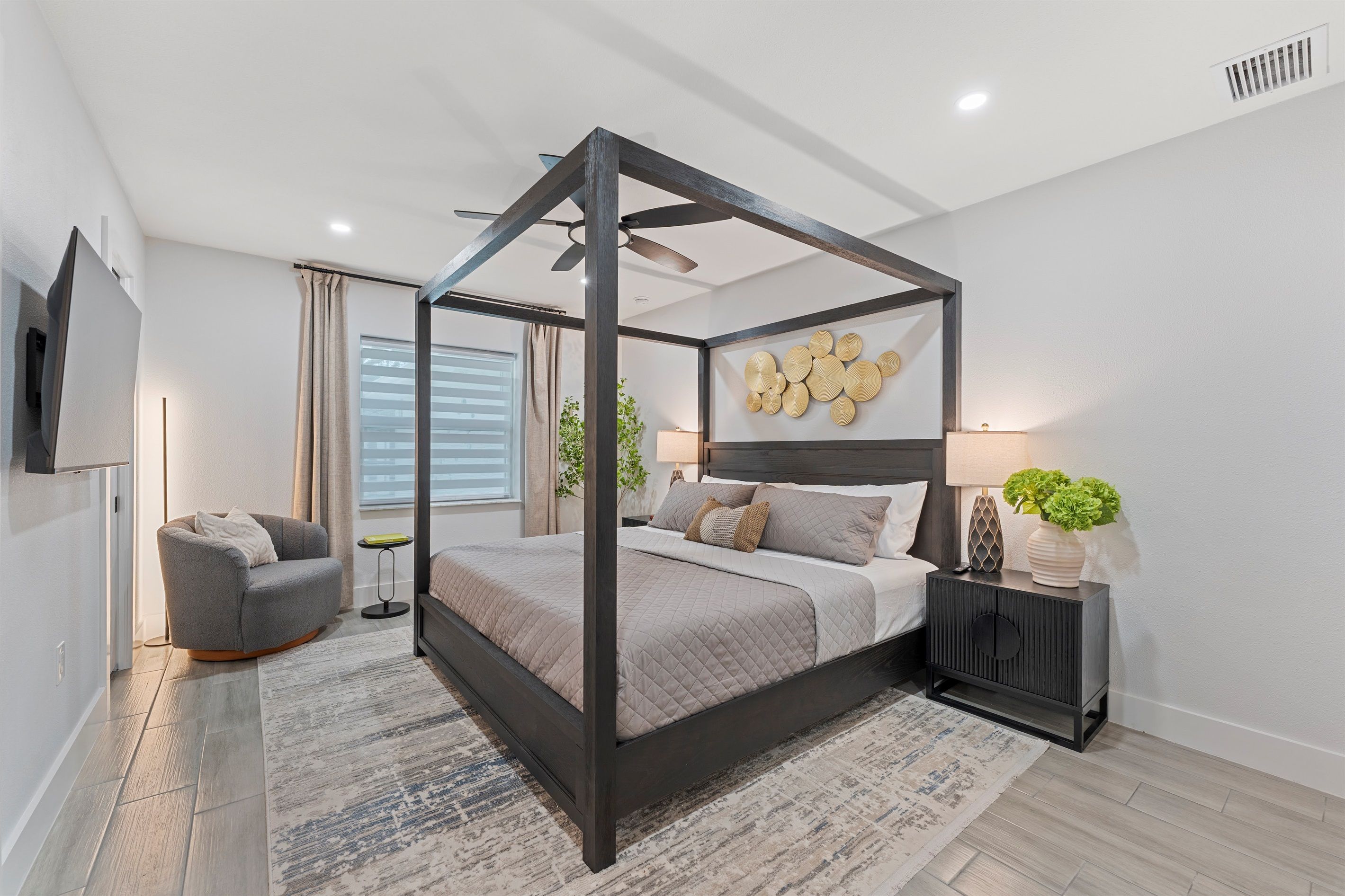 A modern bedroom with a four-poster bed, a grey armchair, a bedside table with a lamp, and a wall-mounted TV.