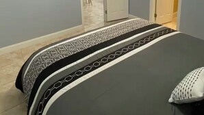 Um quarto com uma cama, um ventilador de teto, uma televisão fixada na parede e uma porta que dá para outro cômodo.