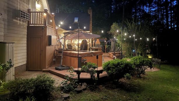 Una terraza de madera con mobiliario exterior y un jacuzzi, rodeada de un jardín y árboles.