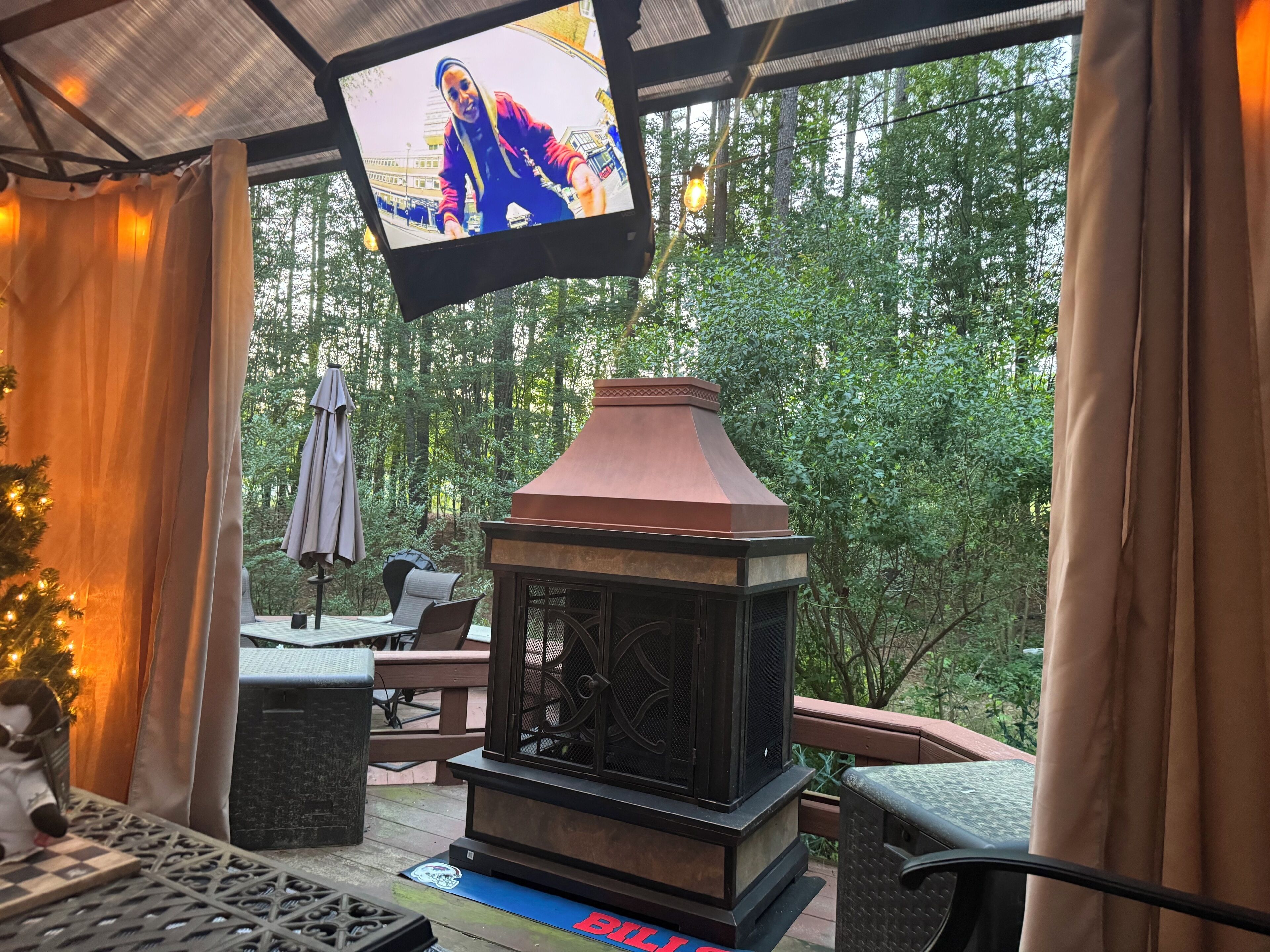 Ein überdachter Außenbereich mit Kamin, Sitzgelegenheiten und einem Fernseher, der eine Person zeigt.