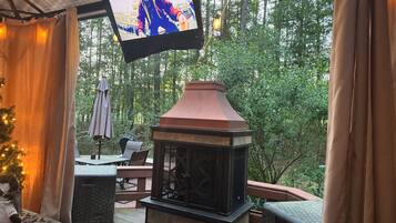Ein überdachter Außenbereich mit Kamin, Sitzgelegenheiten und einem Fernseher, der eine Person zeigt.