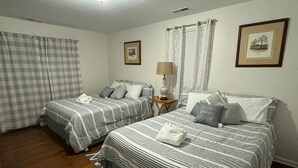 A room with two beds, a wooden floor, a ceiling light, a small table with a lamp, and two framed pictures on the wall.