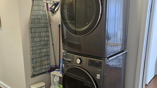 A stacked washer and dryer unit in a laundry room with a mop rack and a small trash bin.