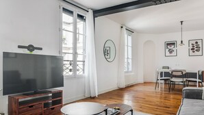 A modern living room with a flat-screen TV, wooden entertainment unit, round coffee table, and a dining area with a white table and chairs.
