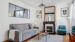 A modern living room with a grey sofa, a small round table, a teal armchair, a fireplace, and a large mirror.