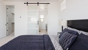 A bedroom with a bed, a nightstand, a lamp, and a sliding door leading to a bathroom. - Modern Desert Home–Great for Families & Groups, Near Golf, Hiking & City Fun (Phoenix)