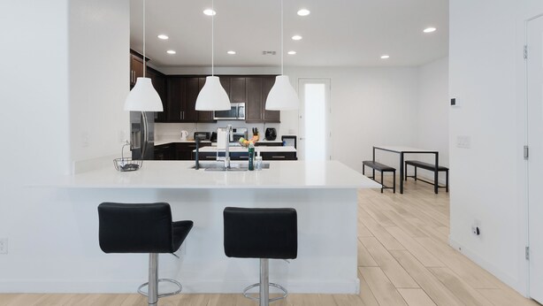 A modern kitchen with white countertops, dark cabinets, and pendant lighting. There are two black bar stools and a dining table with benches in the adjacent area. - Modern Desert Home–Great for Families & Groups, Near Golf, Hiking & City Fun (Phoenix)