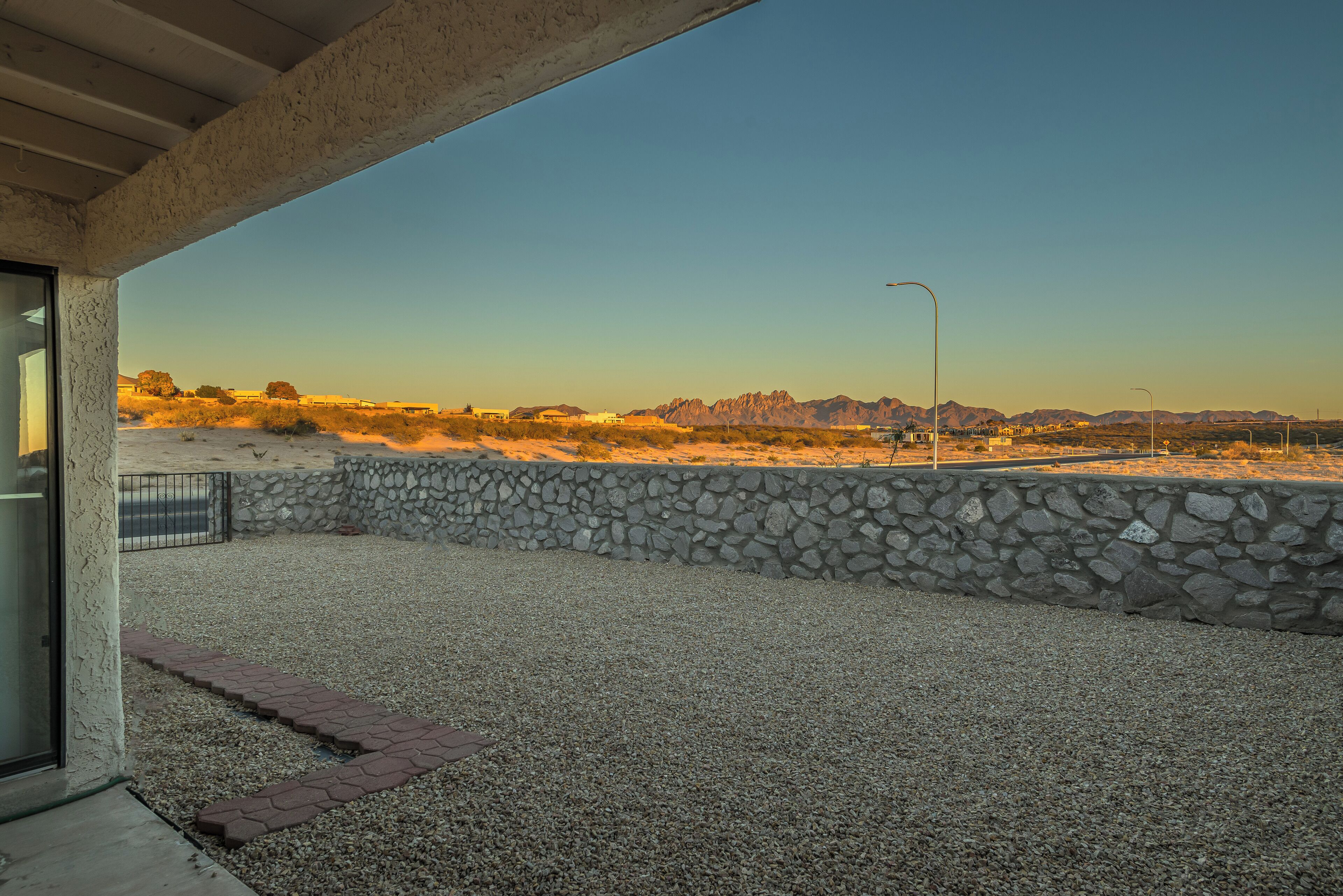 A patio with a stone wall and gravel ground, leading to a view of a desert landscape with mountains in the distance.