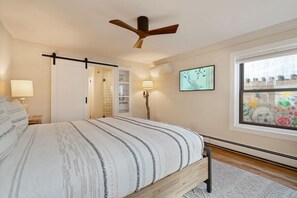 Un dormitorio con cama, ventilador de techo, ventana con vistas y puerta que da a un baño.