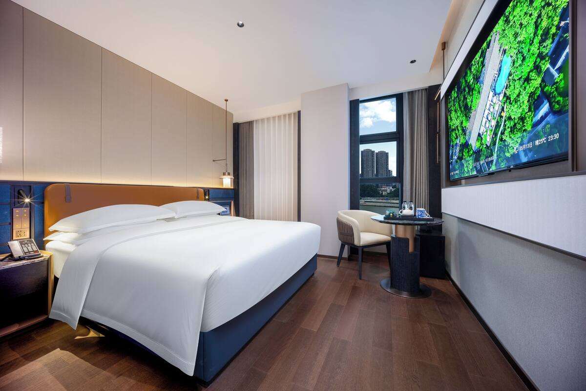 A modern hotel room with a large bed, a desk, and a TV displaying a cityscape.