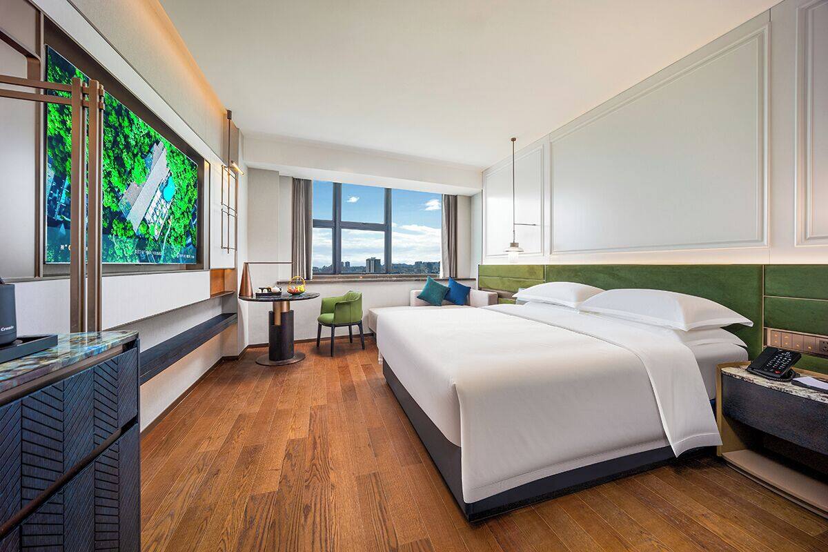 A hotel room with a large bed, a desk, a chair, and a view of the city through the window.