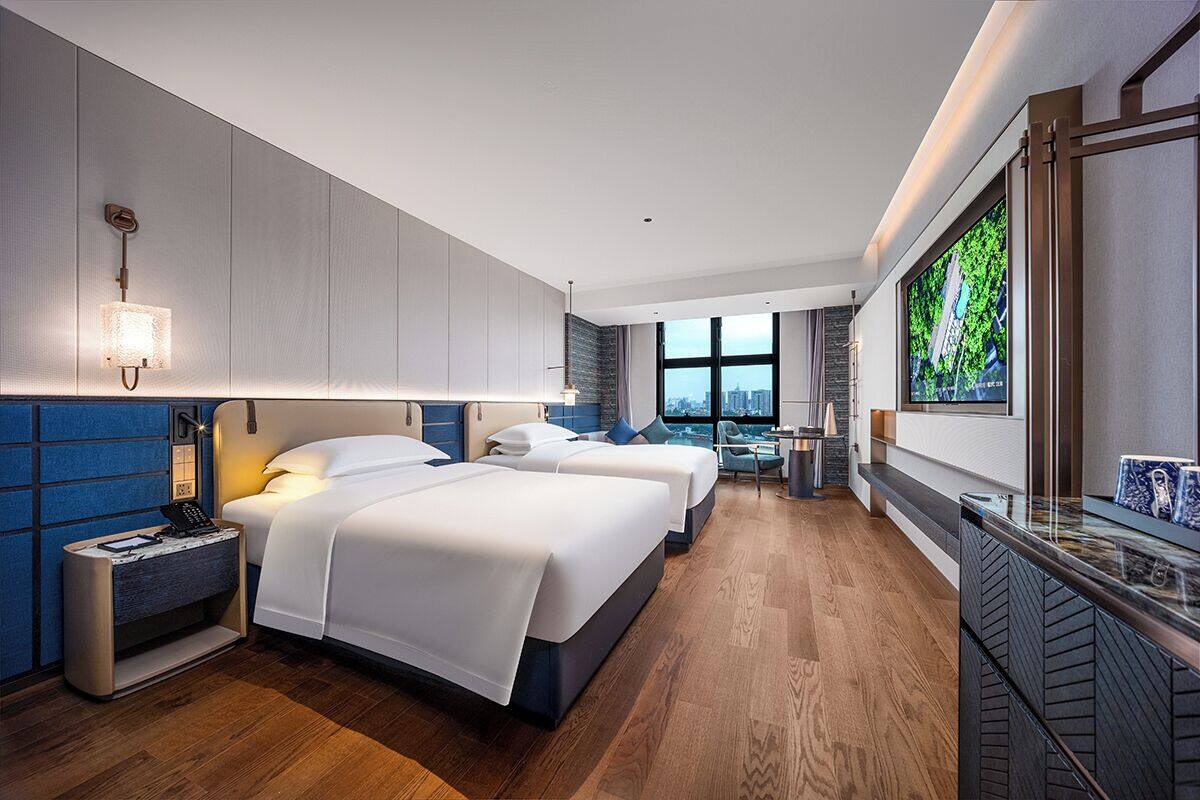 A modern hotel room with two beds, a desk, a TV, and a large window with a city view.