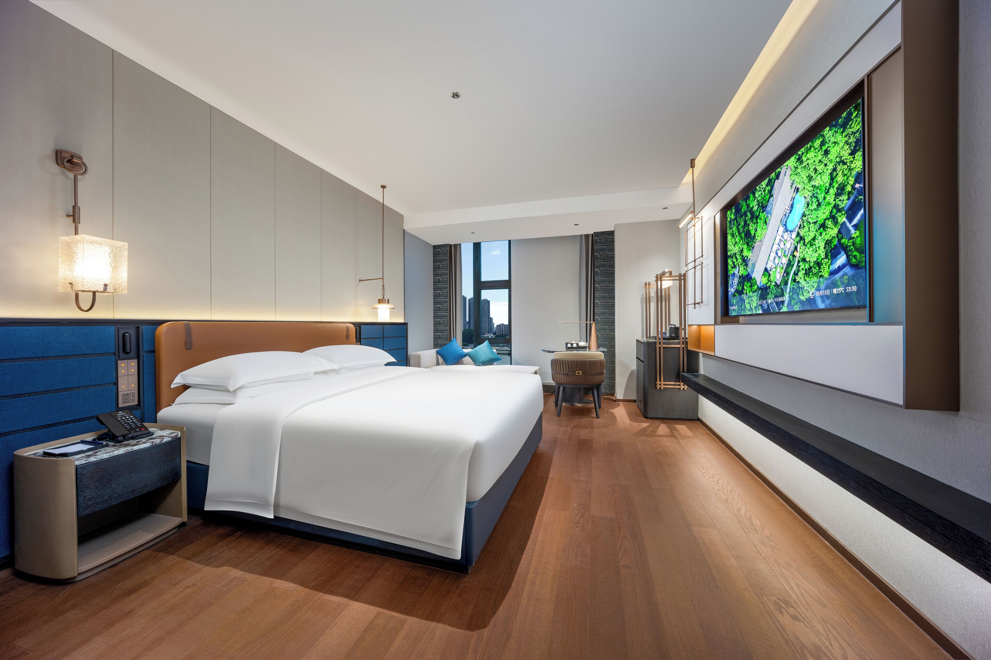 A modern hotel room with a large bed, a desk with a lamp, a TV mounted on the wall, and a view of the city through the window.