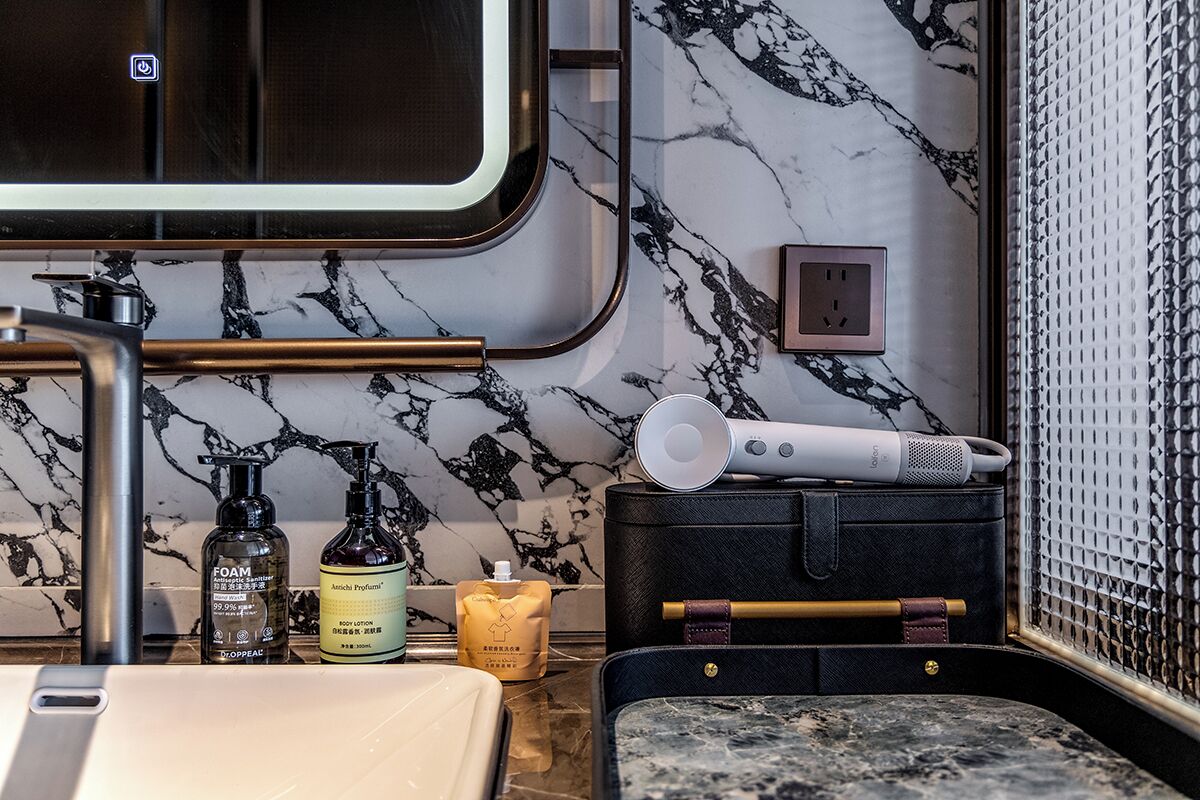 A bathroom with a marble backsplash, a sink, a mirror, and toiletries including a bottle of foam, a body lotion, and a candle.