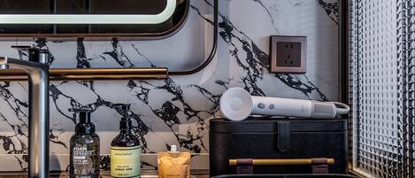 A bathroom with a marble backsplash, a sink, a mirror, and toiletries including a bottle of foam, a body lotion, and a candle.