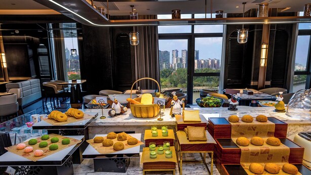 A modern dining area with a variety of pastries and desserts displayed on tables, a chef figurine, and a city view through large windows. - Guangzhou Elegant International Hotel Panyu Square Metro Station Branch - Free Shuttle Bus to Canton Fair Complex During Canton Fair Period & Free Deliver Service to Guangzhou Baiyun International Airport (Guangzhou)