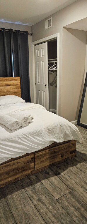 A neatly made bed with white linens and rolled towels, a wooden headboard, and a wooden floor.