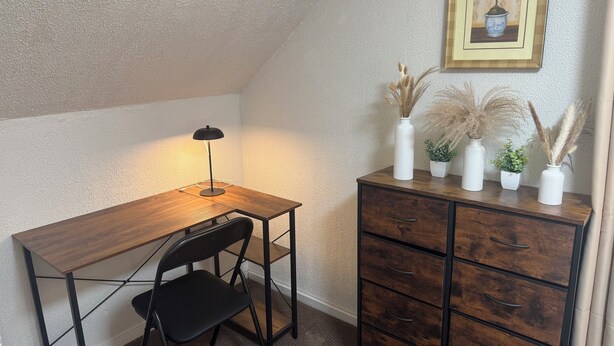 Ein Eckzimmer mit Schreibtisch aus Holz, einem schwarzen Stuhl, einer Schreibtischlampe, einer Kommode mit Schubladen und einem gerahmten Bild an der Wand.