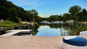 A lakeside dock with a sandy beach, a blue and white floating lounge, and a calm water body surrounded by greenery.