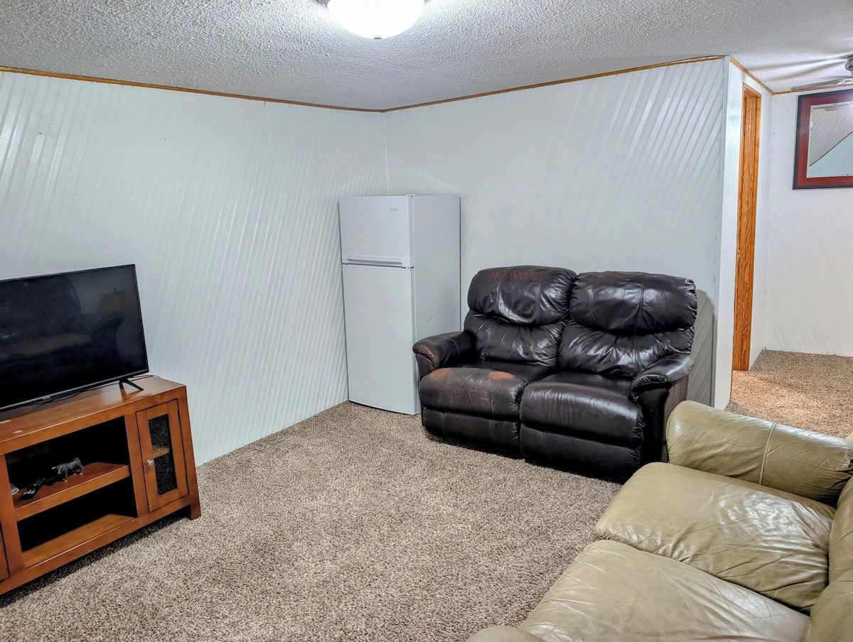A living room with a flat-screen TV on a wooden stand, a leather recliner, a beige sofa, and a white refrigerator.