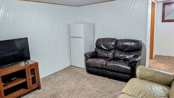 A living room with a flat-screen TV on a wooden stand, a leather recliner, a beige sofa, and a white refrigerator.