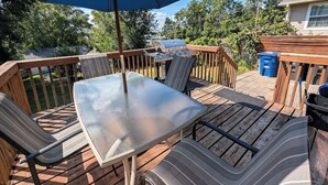 Une terrasse extérieure avec une table à plateau en verre, des chaises rembourrées à rayures, un barbecue et un parasol bleu.