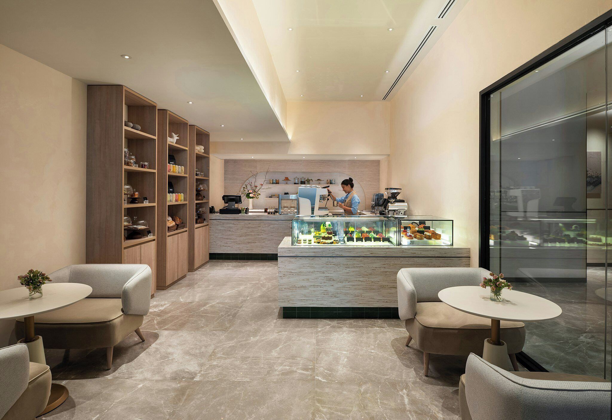 A modern interior with a central counter displaying pastries, flanked by seating areas and wooden shelving units.