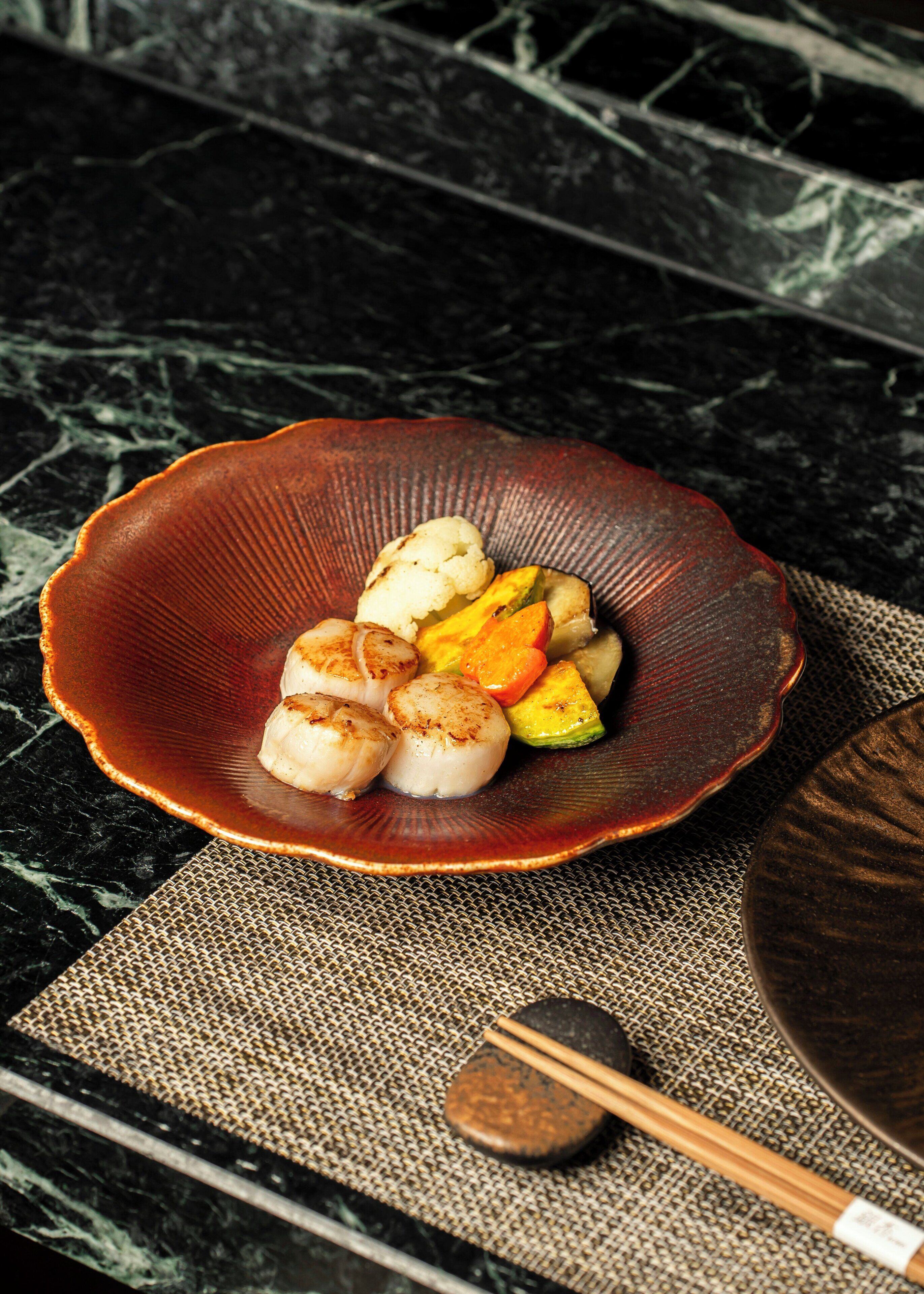 A ceramic bowl with scallops and vegetables on a textured mat.