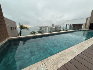 A modern infinity pool with a city view, a wooden deck, and a glass railing. - New, Quiet Apartment with Stunning Ocean View (Lima)