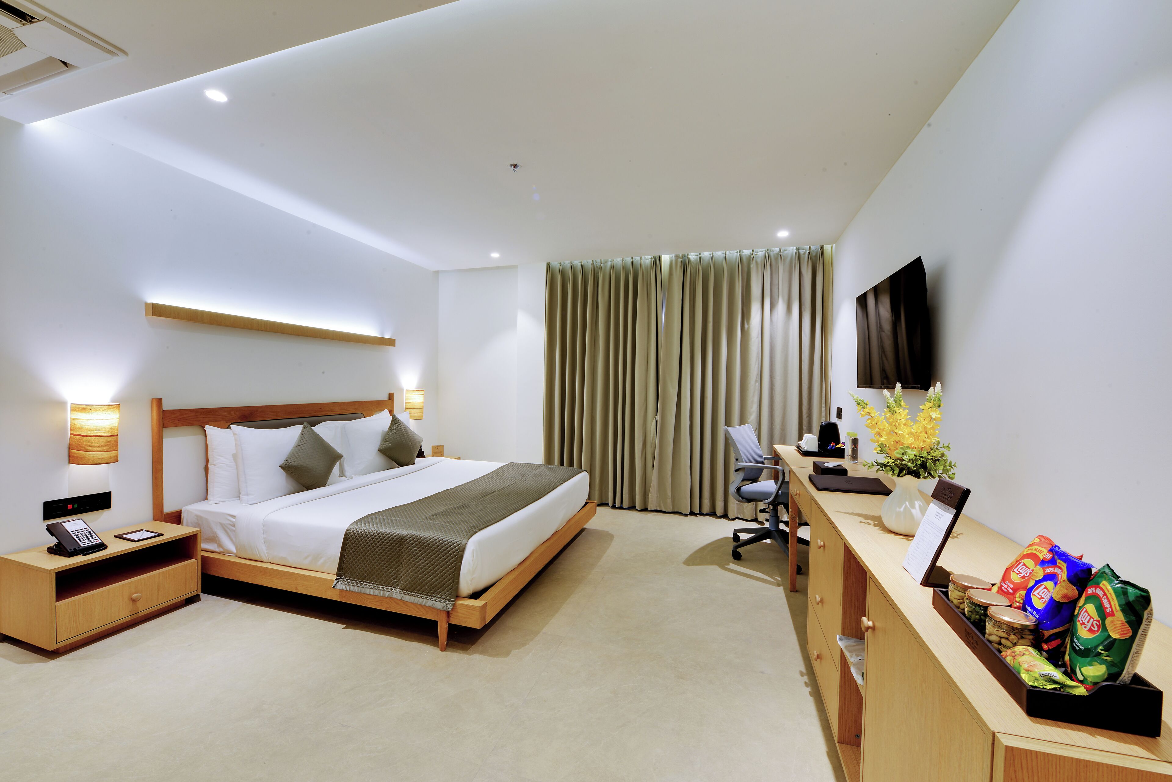 A hotel room with a bed, bedside table, lamp, desk with a computer, and a TV.