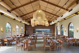 A spacious dining area with multiple tables and chairs, a large chandelier, and a kitchen in the background.