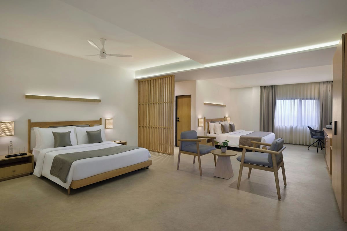 A modern hotel room with a large bed, a desk with a chair, a small table with a plant, and a separate seating area with two chairs.
