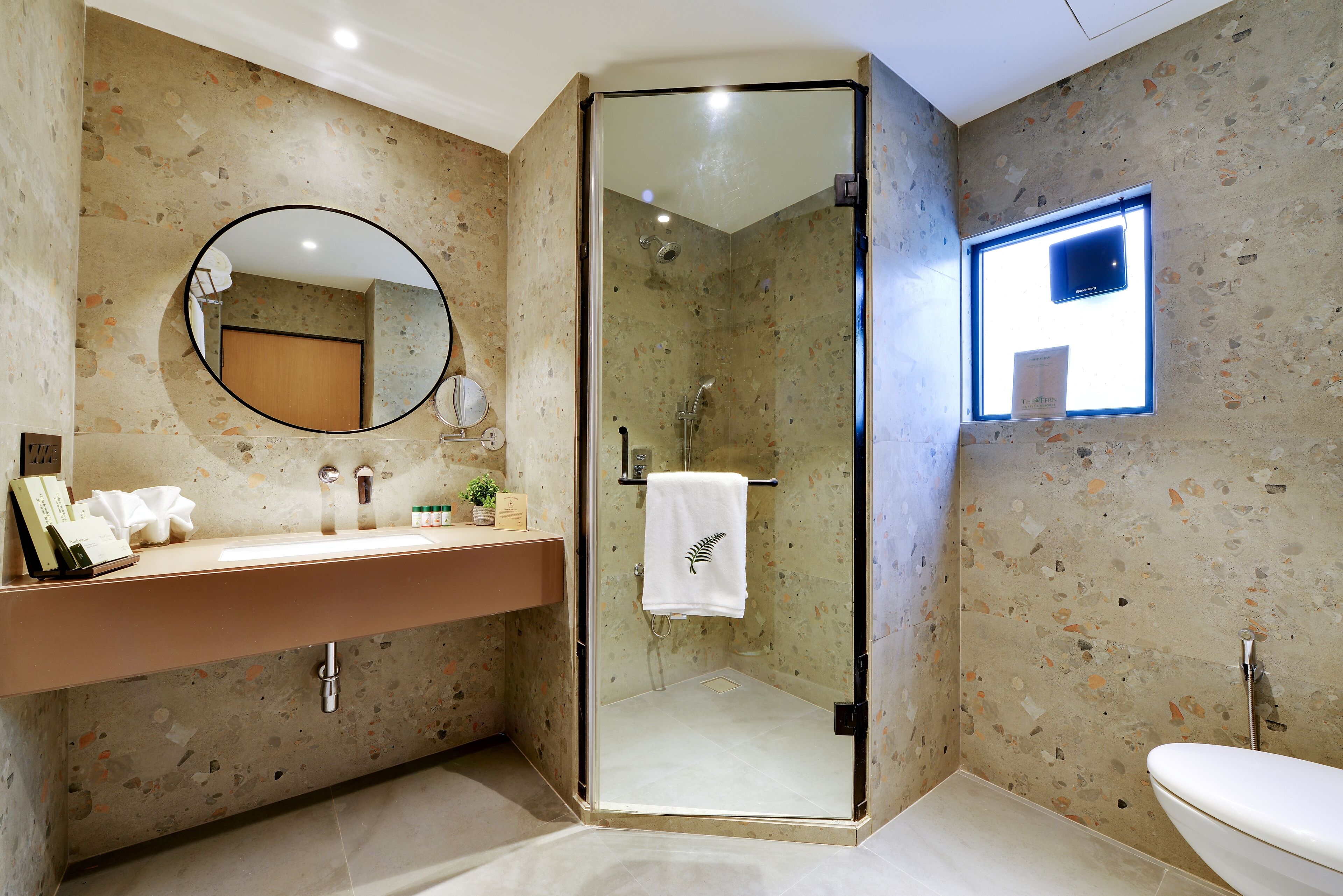 A bathroom with a glass-enclosed shower, a round mirror, a sink with toiletries, and a toilet.