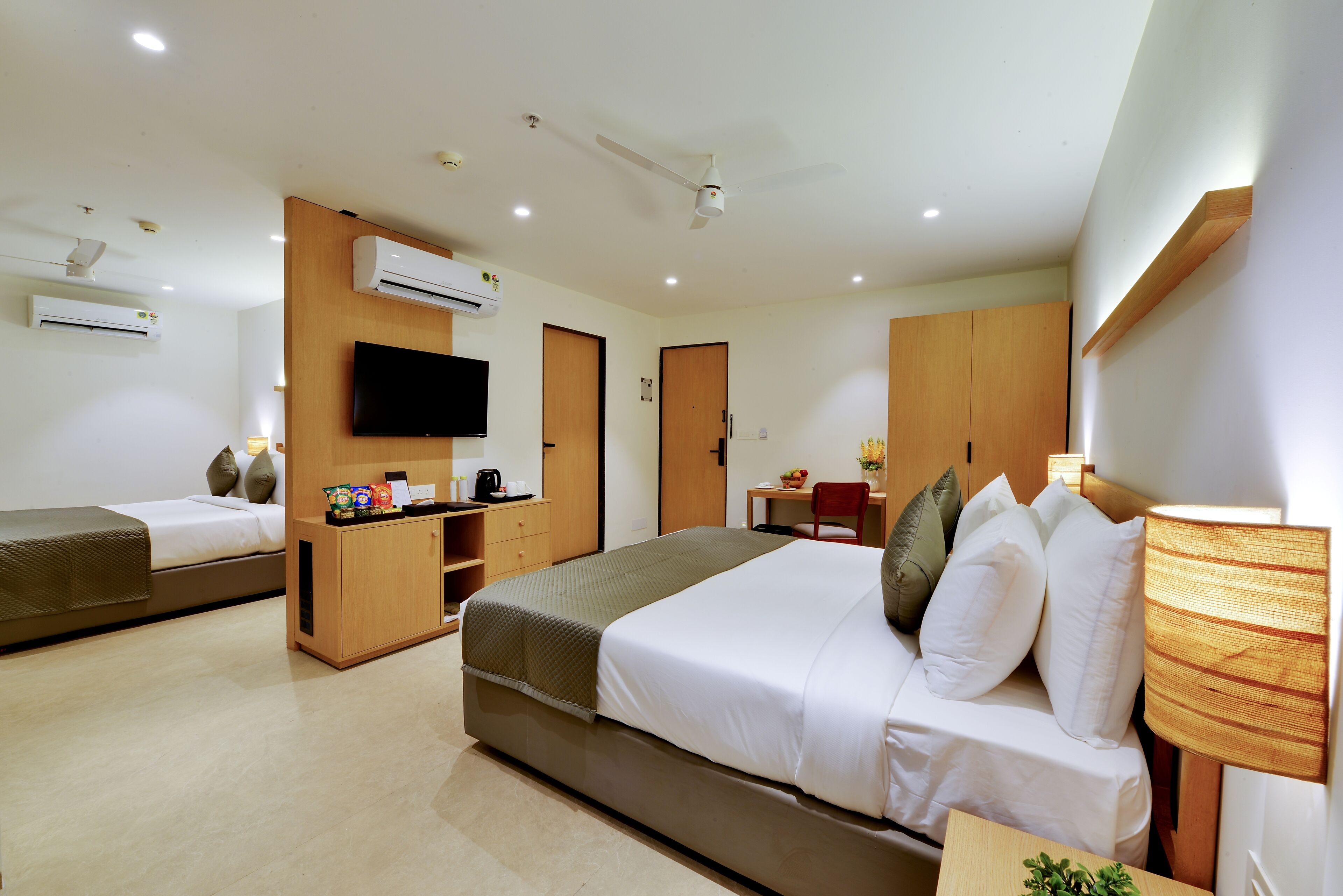 A hotel room with two beds, a TV, a desk with a chair, and a small table with a lamp.