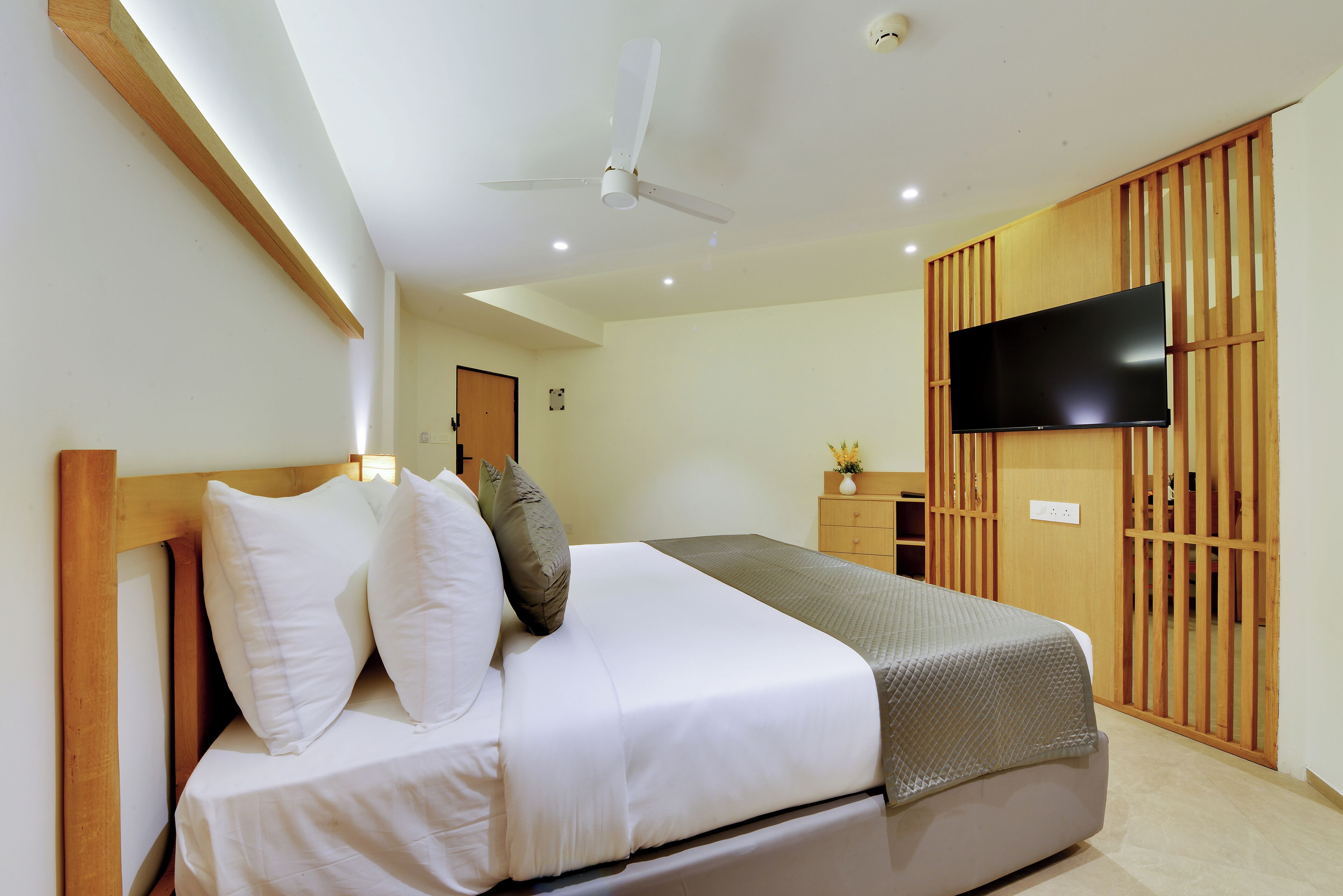 A neatly made bed with pillows, a ceiling fan, a flat-screen TV mounted on a wooden panel, and a wooden wardrobe with a sliding door.