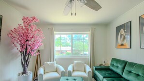 Ein Wohnzimmer mit einem grünen Sofa, zwei weißen Sesseln, einem grünen Hocker, einem großen Fenster mit Vorhängen, einem Deckenventilator und einem dekorativen rosa Baum.